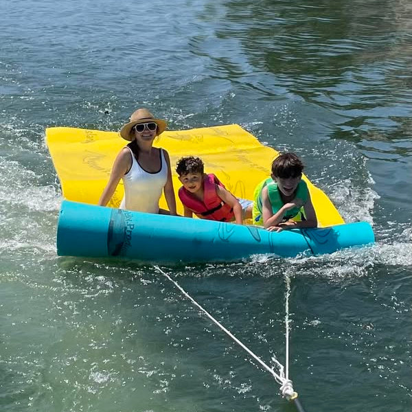 Family Being Towed behind a Pontoon boat on thier floating foam mat by Aqua Lily Pad. This pad has the Lily Surfer Kit installed on it to make towing possible.