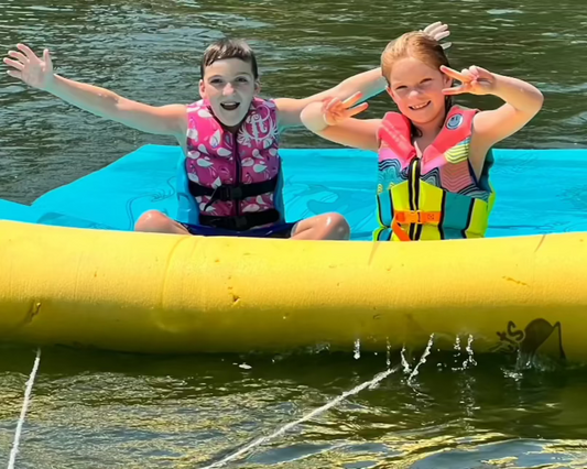 Two kids sitting on a lily pad that has a Lily Surfer kit installed so their parents can tube them around the lake behind their boat. Kids are smiling and look like they are having a blast.