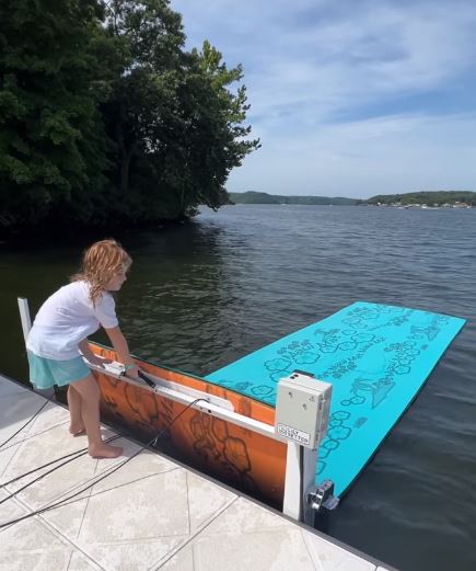 A young red head girl lifts a big floating mat lily pad out of the water using a unique device called a dock setter which installs on the side of dock to help lift , roll, strap and store large floating mats used for recreation