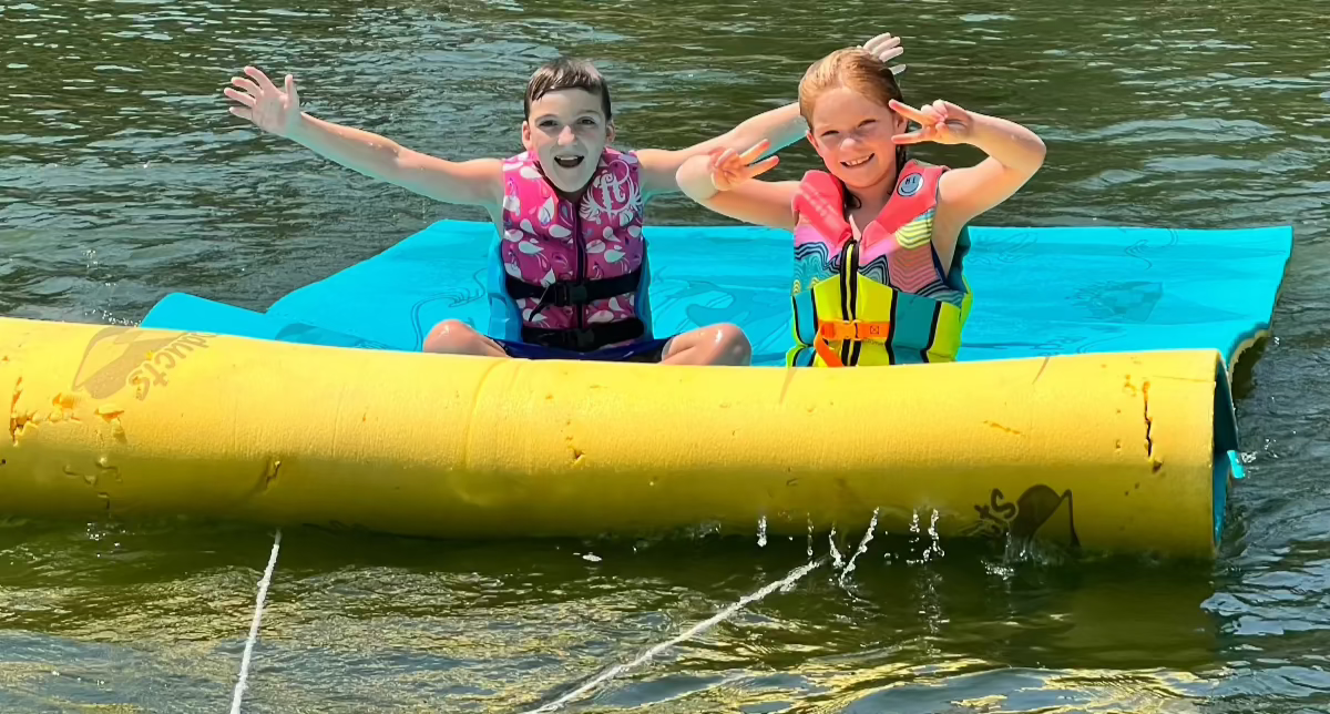 Two kids sitting on a lily pad that has a Lily Surfer kit installed so their parents can tube them around the lake behind their boat.   Kids are smiling and look like they are having a blast. 
